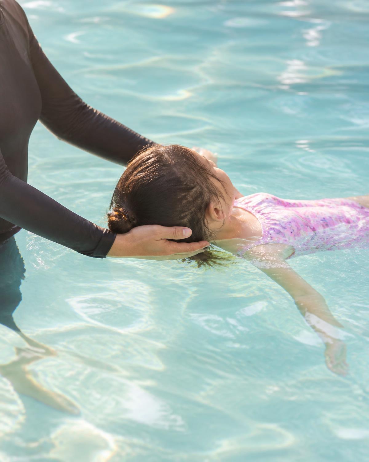 Instructor's hands gently supporting a child learning to float in a sunlit pool